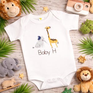 May include: A white short-sleeved baby bodysuit featuring an embroidered design of a gray elephant and a yellow giraffe, with the text "Baby H" printed in brown below the animals. The garment has a round neckline and snap closures at the bottom.