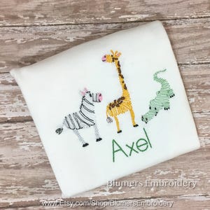 May include: A white shirt featuring embroidered designs of a zebra with black stripes, a yellow giraffe with brown spots, and a green alligator. The name Axel is embroidered in green thread below the animals. The text Blumers Embroidery is visible.