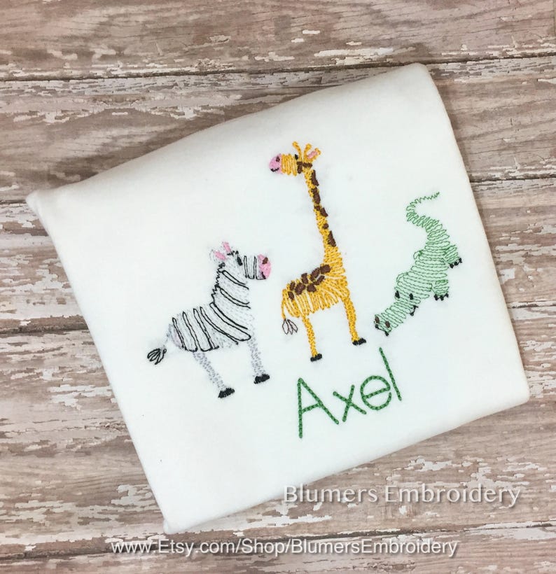 May include: A white shirt featuring embroidered designs of a zebra with black stripes, a yellow giraffe with brown spots, and a green alligator. The name Axel is embroidered in green thread below the animals. The text Blumers Embroidery is visible.