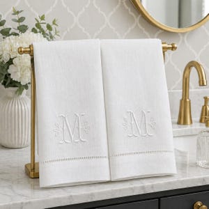 May include: Two white rectangular hand towels draped over a gold metal stand. Each towel features a centered white embroidered letter M with decorative flourishes and a horizontal row of small cutwork detailing near the bottom edge.