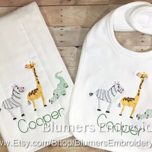 May include: A white burp cloth and a matching white baby bib, both featuring embroidered designs of a zebra, a giraffe, and a crocodile. The name Cooper is stitched in green thread below the animals on both items. The text Blumers Embroidery is visible.