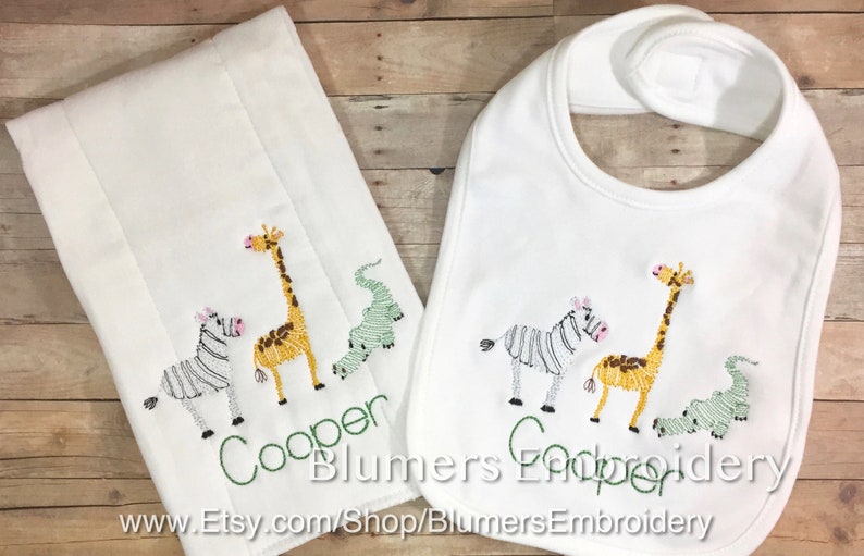 May include: A white burp cloth and a matching white baby bib, both featuring embroidered designs of a zebra, a giraffe, and a crocodile. The name Cooper is stitched in green thread below the animals on both items. The text Blumers Embroidery is visible.