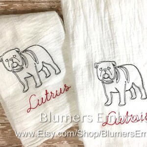 K&ouml;nnte beinhalten: Zwei wei&szlig;e K&uuml;chent&uuml;cher mit einer schwarzen Stickerei, die den Umriss einer Bulldogge zeigt. Unter jedem Hund ist der Name Lutrus in rotem Schreibschriftgarn gestickt. Die T&uuml;cher haben eine strukturierte Oberfl&auml;che und liegen nebeneinander.
