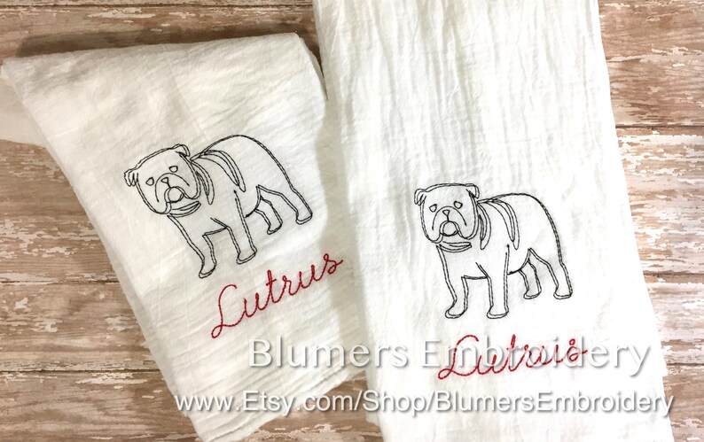 K&ouml;nnte beinhalten: Zwei wei&szlig;e K&uuml;chent&uuml;cher mit einer schwarzen Stickerei, die den Umriss einer Bulldogge zeigt. Unter jedem Hund ist der Name Lutrus in rotem Schreibschriftgarn gestickt. Die T&uuml;cher haben eine strukturierte Oberfl&auml;che und liegen nebeneinander.