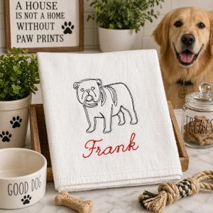 K&ouml;nnte beinhalten: Ein wei&szlig;es K&uuml;chentuch mit einer schwarzen Stickerei, die den Umriss einer Bulldogge zeigt, sowie dem Namen Frank in rotem Garn. Das Tuch ist gefaltet auf einem Holzst&auml;nder drapiert und pr&auml;sentiert ein klares Design mit detaillierter Stickerei.