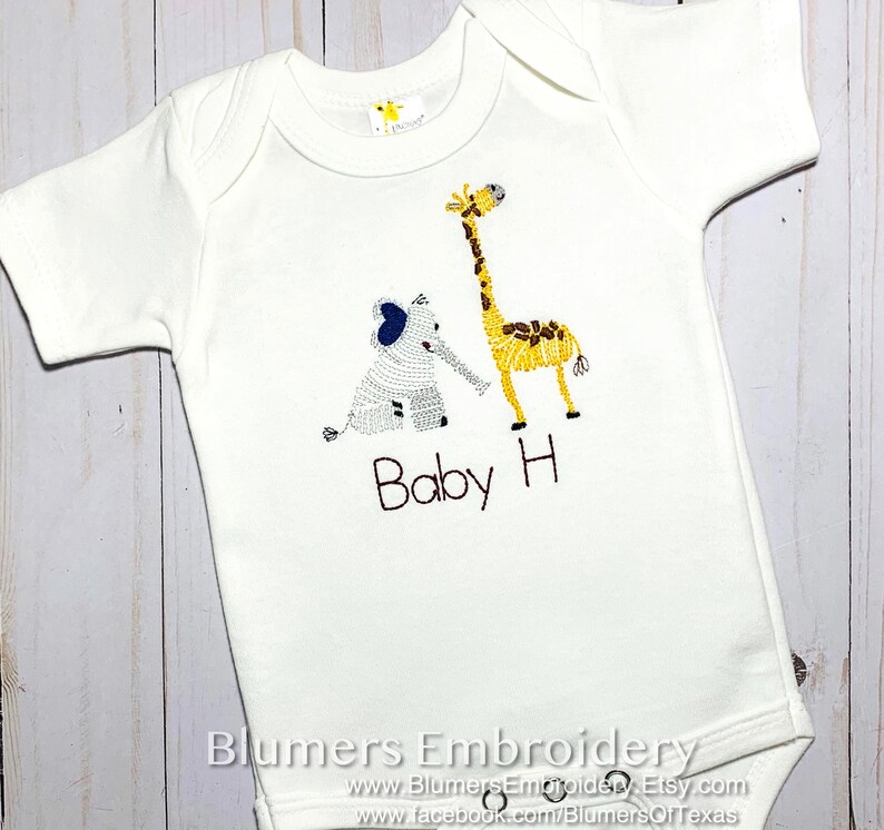 May include: A white short-sleeved baby bodysuit featuring an embroidered gray elephant and a yellow giraffe. Below the animals, the text Baby H is embroidered in black thread. The garment has a round neckline and three metal snaps at the bottom.