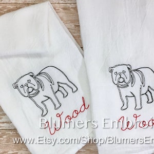 K&ouml;nnte beinhalten: Zwei wei&szlig;e K&uuml;chent&uuml;cher mit schwarzen, gestickten Umrissen einer Bulldogge. Unter jedem Hund ist der Name Wood in roter Schreibschrift eingestickt. Die T&uuml;cher sind gefaltet und nebeneinander angeordnet, was die detaillierte Stickerei zeigt.