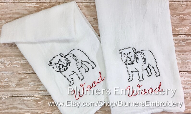 K&ouml;nnte beinhalten: Zwei wei&szlig;e K&uuml;chent&uuml;cher mit schwarzen, gestickten Umrissen einer Bulldogge. Unter jedem Hund ist der Name Wood in roter Schreibschrift eingestickt. Die T&uuml;cher sind gefaltet und nebeneinander angeordnet, was die detaillierte Stickerei zeigt.