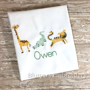 May include: A white fabric item featuring embroidered designs of a yellow giraffe, a light green alligator, and a yellow tiger with black stripes. The name Owen is embroidered in green thread below the animals. The text Blumers Embroidery is visible.