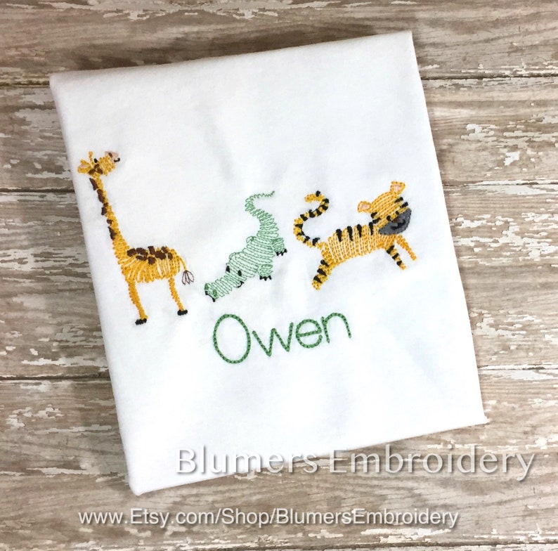 May include: A white fabric item featuring embroidered designs of a yellow giraffe, a light green alligator, and a yellow tiger with black stripes. The name Owen is embroidered in green thread below the animals. The text Blumers Embroidery is visible.
