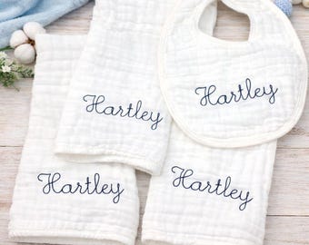 Könnte beinhalten: Ein weißes Baby-Geschenkset mit einem Lätzchen und drei Waschlappen. Jeder Artikel ist mit dem Namen "Hartley" in marineblauer Schrift bestickt. Das Set besteht aus einem weichen, strukturierten Stoff, ideal für ein Baby.