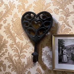 May include: A black, heart-shaped wall hook with an ornate, openwork design. The hook is mounted on a wall with a floral pattern wallpaper. A framed black and white picture hangs nearby.