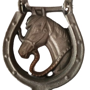 May include: A cast iron door knocker shaped like a horseshoe with a horse's head in the center. The horseshoe is a dark gray color and the horse's head is a lighter gray color.