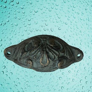 May include: A dark, ornate metal drawer pull with a decorative floral design in gold accents. The pull has a curved shape with two holes for mounting. The finish appears aged, with a distressed look.