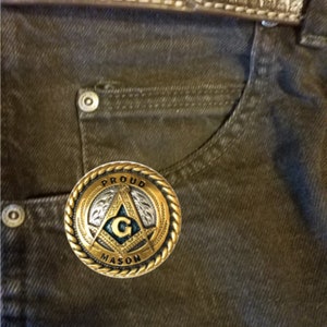 May include: A gold-colored belt buckle with a Masonic symbol and the words "Proud Mason" around the edge. The buckle is on a pair of brown jeans.