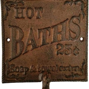 May include: A rectangular, cast iron sign with the words "HOT BATHS" in large, embossed letters. The sign is a rusty brown color and includes the text "25¢" and "Soap & towel extra". A small hook is attached at the bottom.