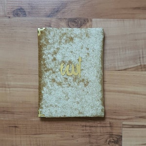 May include: A rectangular, gold-coloured book cover with a plush, textured surface. The cover features a gold-coloured Arabic calligraphy design. The corners are reinforced with gold-coloured metal. The book is resting on a wooden surface.
