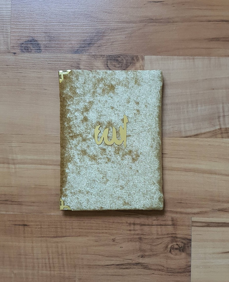 May include: A rectangular, gold-coloured book cover with a plush, textured surface. The cover features a gold-coloured Arabic calligraphy design. The corners are reinforced with gold-coloured metal. The book is resting on a wooden surface.