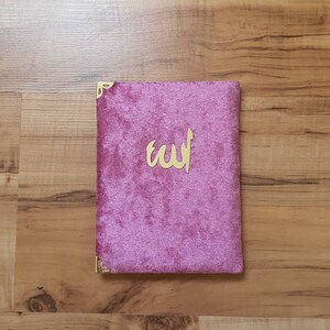 May include: A pink velvet book cover with gold corners and the word "الله" in gold lettering.
