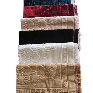 Op de afbeelding: Een stapel gebedskleden in verschillende kleuren, waaronder marineblauw, bordeauxrood, beige, zwart, wit en bruin. Elk kleed heeft een reliëf ontwerp en gefranjerde randen. De kleden zijn netjes opgevouwen en op elkaar gestapeld.