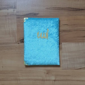 May include: A turquoise velvet book cover with gold corner protectors and gold Arabic calligraphy. The cover is rectangular and has a soft, plush texture. The Arabic text is centered on the cover.