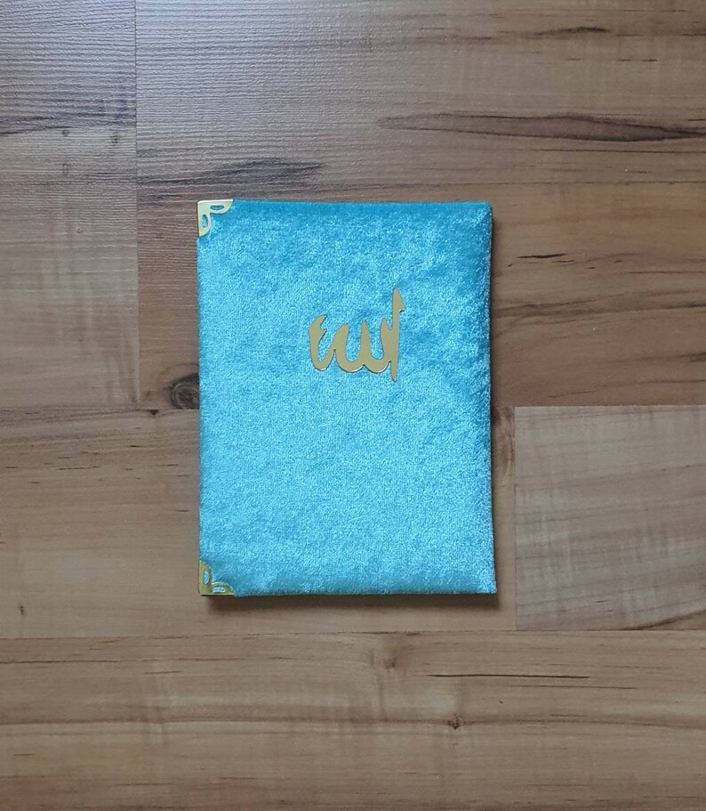 May include: A turquoise velvet book cover with gold corner protectors and gold Arabic calligraphy. The cover is rectangular and has a soft, plush texture. The Arabic text is centered on the cover.
