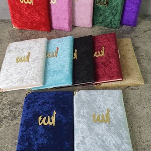 May include: A selection of velvet-covered books in colours such as red, pink, green, and blue. Each book has gold-coloured Arabic script on the cover and gold corner protectors. The books are displayed on a grey surface.