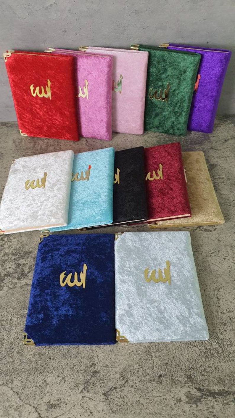 May include: A selection of velvet-covered books in colours such as red, pink, green, and blue. Each book has gold-coloured Arabic script on the cover and gold corner protectors. The books are displayed on a grey surface.