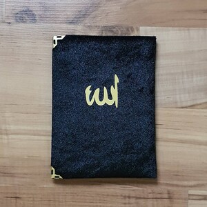May include: A black velvet book cover with gold corner protectors and gold Arabic calligraphy. The cover is rectangular and sits on a wooden surface.