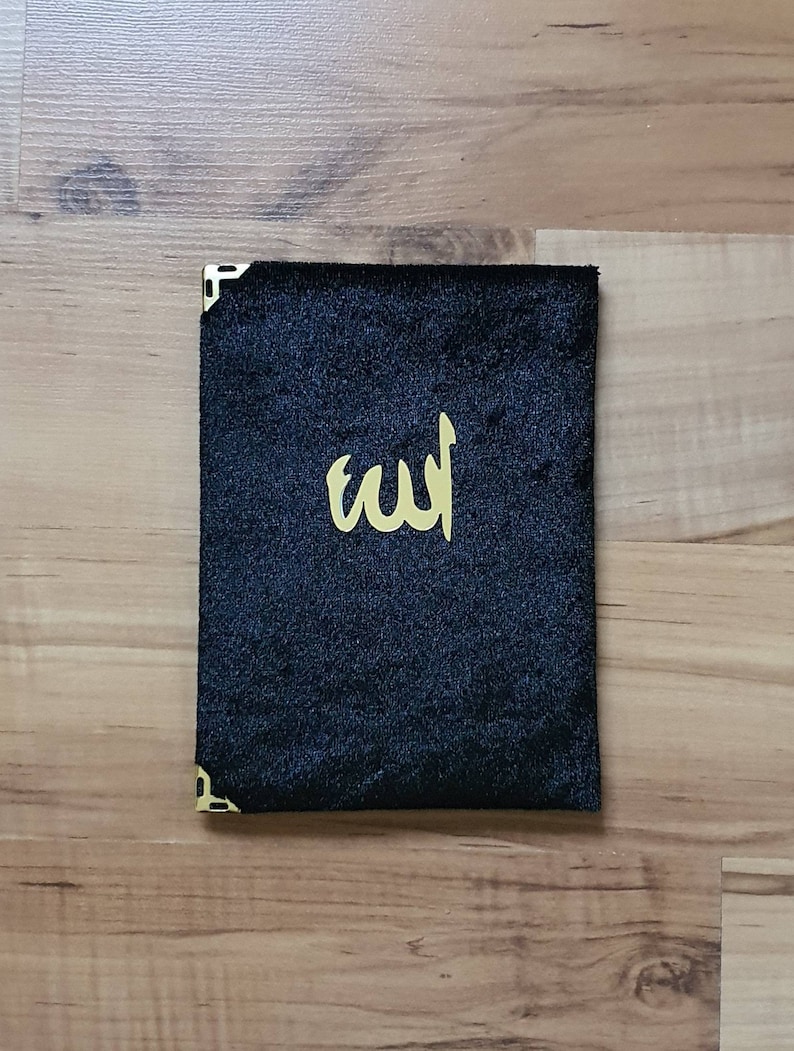 May include: A black velvet book cover with gold corner protectors and gold Arabic calligraphy. The cover is rectangular and sits on a wooden surface.