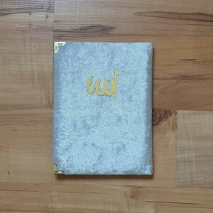 May include: A light blue velvet book with gold corner protectors and gold Arabic calligraphy. The book is lying on a wooden surface.