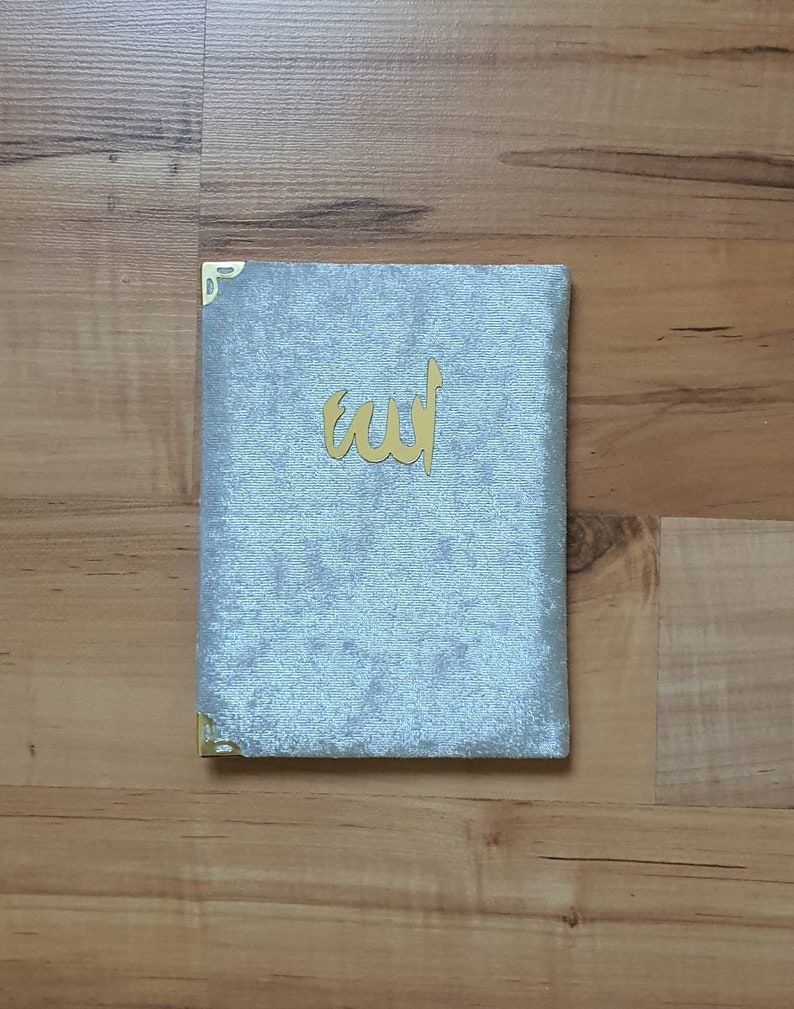 May include: A light blue velvet book with gold corner protectors and gold Arabic calligraphy. The book is lying on a wooden surface.