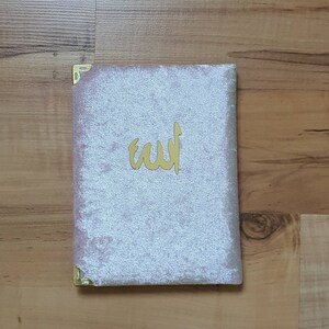 May include: A light pink velvet book cover with gold corner protectors and a gold Arabic inscription. The book is rectangular and sits on a wooden surface.