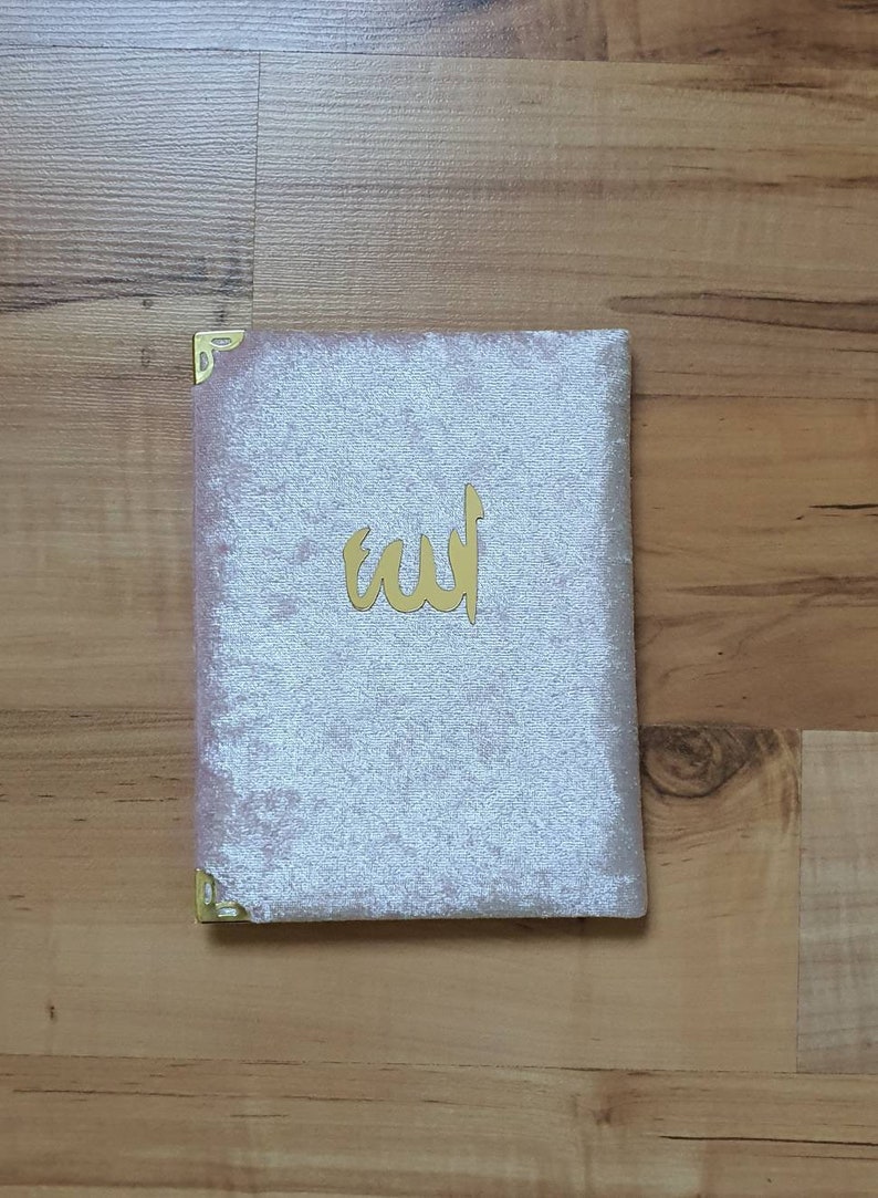 May include: A light pink velvet book cover with gold corner protectors and a gold Arabic inscription. The book is rectangular and sits on a wooden surface.
