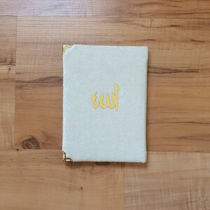 May include: A white velvet book cover with gold corners and the word "Allah" written in gold lettering.