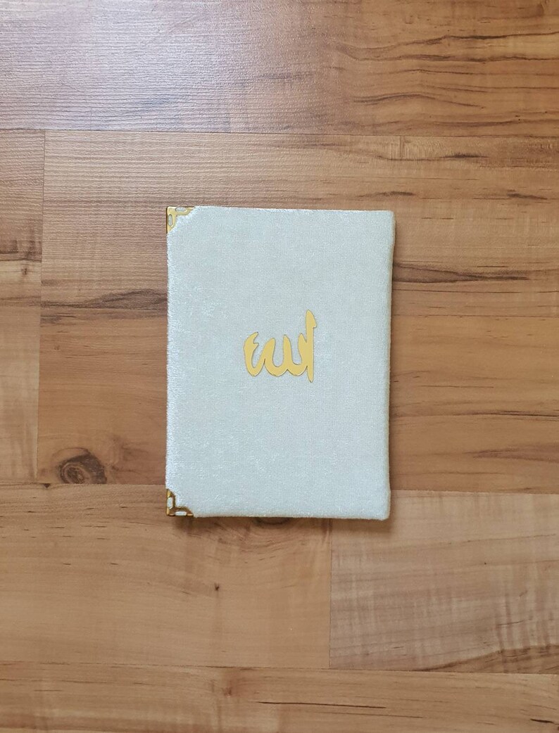 May include: A white velvet book cover with gold corners and the word "Allah" written in gold lettering.