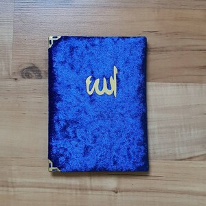 May include: A blue velvet book cover with gold corner accents and gold Arabic calligraphy. The cover is a deep blue colour and has a soft, textured appearance. The gold calligraphy is centred on the cover.