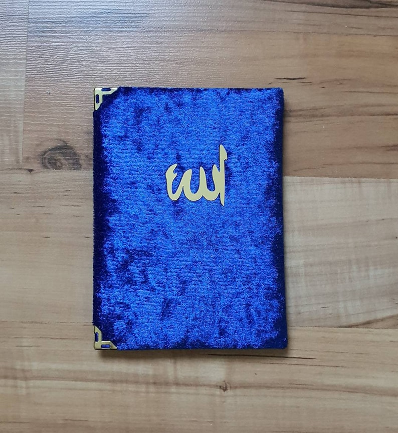 May include: A blue velvet book cover with gold corner accents and gold Arabic calligraphy. The cover is a deep blue colour and has a soft, textured appearance. The gold calligraphy is centred on the cover.