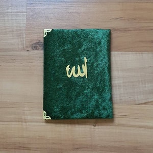 May include: A dark green velvet book cover with gold corner detailing and gold Arabic script. The cover is rectangular and has a soft, plush texture. The calligraphy is centered on the cover.
