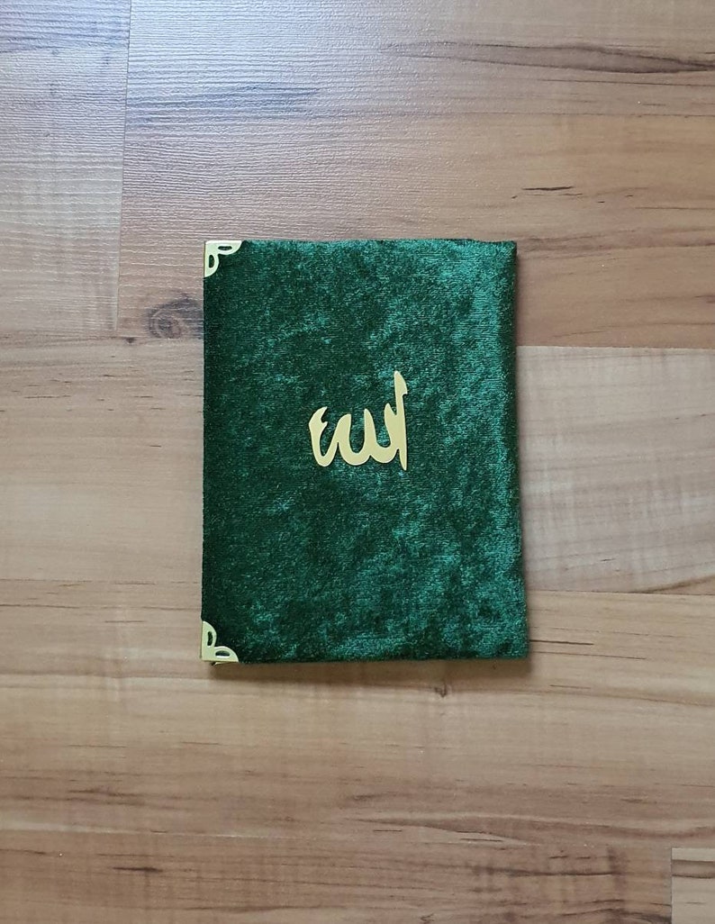 May include: A dark green velvet book cover with gold corner detailing and gold Arabic script. The cover is rectangular and has a soft, plush texture. The calligraphy is centered on the cover.