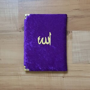 May include: A deep purple velvet book cover with gold corner protectors and gold Arabic calligraphy. The cover has a soft texture. The calligraphy reads "Allah".