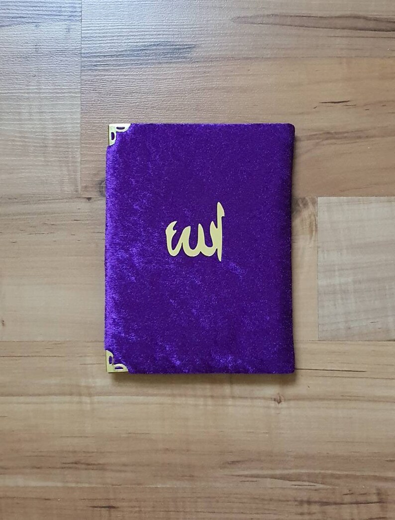 May include: A deep purple velvet book cover with gold corner protectors and gold Arabic calligraphy. The cover has a soft texture. The calligraphy reads "Allah".