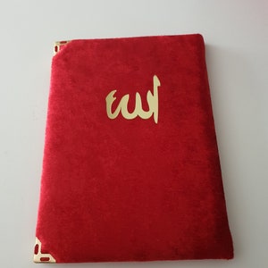 May include: A red velvet book with gold corner detailing and gold Arabic script. The cover displays the word "Allah" in gold. The book is rectangular and appears closed.