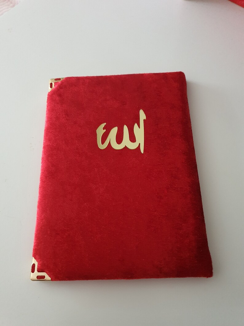 May include: A red velvet book with gold corner detailing and gold Arabic script. The cover displays the word "Allah" in gold. The book is rectangular and appears closed.