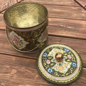 Könnte beinhalten: Eine offene, vintage Metallbox mit Deckel. Die Box ist olivgrün mit floralen und goldenen Akzenten. Der Deckel liegt daneben und gibt den Blick ins Innere frei. Der Griff ist goldfarben. Das Ganze steht auf einer Holzoberfläche.