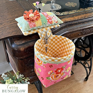 May include: A fabric pincushion with a floral top and a matching hanging storage basket featuring a pink floral exterior and yellow gingham interior. The set includes a pair of metal scissors and three pins. The text Curry BUNGALOW is visible.