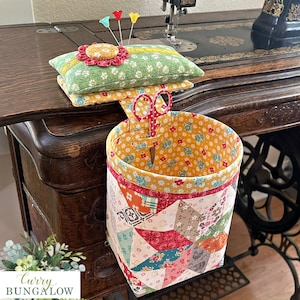 May include: A fabric pincushion with a floral green top and a yellow flower accent, attached to a hanging cylindrical fabric basket featuring a patchwork design of various colorful patterns, including a pair of red scissors with white dots.