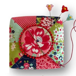 May include: A patchwork fabric pincushion with a red, pink, green, and blue floral design. The pincushion has a round center with a red and pink floral design. Three colorful flower-shaped pins are stuck in the pincushion. A pair of red scissors with white polka dots is next to the pincushion. The text "Curry Bungalow" is written below the pincushion.