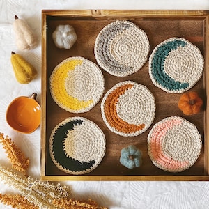 May include: Six crocheted coasters in a wooden tray. The coasters are beige with different coloured crescent moons. The colours are yellow, orange, green, pink, black, and grey.