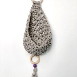 May include: A gray crocheted hanging basket with a wooden ring at the top and a wooden ring with a purple bead and tassel at the bottom.